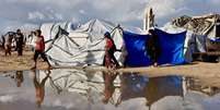 Palestinos de Gaza caminham ao lado de uma grande po&ccedil;a de &aacute;gua perto de seus abrigos, em 28 de dezembro de 2025. Regi&atilde;o enfrenta condi&ccedil;&otilde;es clim&aacute;ticas severas de inverno.  Foto: AFP - OMAR AL-QATTAA / RFI