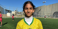 Uma menina palestina olha para a câmera. Ela está em um campo de futebol e veste a camisa da seleção brasileira  Foto: BBC News Brasil