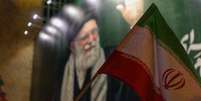 Uma bandeira nacional iraniana &eacute; hasteada em frente a uma faixa com a imagem de Ali Khamenei durante uma vig&iacute;lia em mem&oacute;ria do l&iacute;der supremo do Ir&atilde;, Ali Khamenei, ap&oacute;s a m&iacute;dia estatal iraniana confirmar sua morte em 1&ordm; de mar&ccedil;o de 2026, em Teer&atilde;, Ir&atilde;. Foto: Majid Saeedi/Getty Images