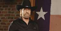 Chuck Norris usa uma camisa preta e um chap&eacute;u de caub&oacute;i enquanto posa para uma foto. Atr&aacute;s dele, v&ecirc;-se uma bandeira do Texas e uma parede de tijolos. Foto: Getty Images / BBC News Brasil