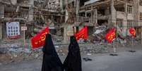 Duas mulheres vestidas de preto dos p&eacute;s &agrave; cabe&ccedil;a caminham diante de pr&eacute;dios destru&iacute;dos e uma s&eacute;rie de bandeiras vermelhas com inscri&ccedil;&otilde;es em persa em amarelo.  Foto: Getty Images / BBC News Brasil