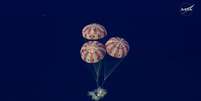 Imagem a&eacute;rea mostra tr&ecirc;s paraquedas com listras brancas e vermelhas inflados amortecendo a aterrissagem da c&aacute;psula  Foto: NASA / EPA / BBC News Brasil
