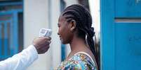 Um m&eacute;dico verifica a temperatura de uma mulher usando um term&ocirc;metro infravermelho sem contato. Somente o bra&ccedil;o do m&eacute;dico, que est&aacute; vestido de branco, &eacute; mostrado. A mulher que est&aacute; com a temperatura verificada tem o cabelo tran&ccedil;ado. Ela est&aacute; vestindo uma blusa azul brilhante com um padr&atilde;o e est&aacute; ao lado de uma porta de madeira azul com um pr&eacute;dio branco e azul ao fundo.  Foto: Getty Images / BBC News Brasil