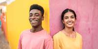 Um homem e uma mulher jovens aparecem lado a lado diante de uma parede pintada com cores vibrantes. Ele veste uma camiseta rosa diante de uma parte amarela da parede, enquanto ela usa uma camiseta amarela diante de uma área rosa  Foto: Getty Images / BBC News Brasil