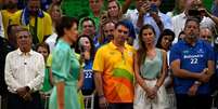 Michelle aparece em primeiro plano, desfocada, em vestido verde e falando ao microfone em um com&iacute;cio eleitoral de 2022. Ao fundo, Fl&aacute;vio e sua esposa observam Michelle ao lado de outras pessoas  Foto: MAURO PIMENTEL/AFP via Getty Images / BBC News Brasil