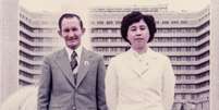 Uma fotografia antiga de Hitomi e seu marido, Charles. Eles est&atilde;o em p&eacute; em frente a uma fonte e um pr&eacute;dio de apartamentos. Hitomi veste um blazer com bot&otilde;es e uma saia. Charles usa um terno cinza com uma gravata listrada. Hitomi est&aacute; com os bra&ccedil;os atr&aacute;s das costas e Charles com os bra&ccedil;os ao lado do corpo. Ambos sorriem e olham diretamente para a c&acirc;mera. Foto: Hitomi Soga / BBC News Brasil