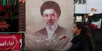 Uma mulher caminha ao lado de um cartaz com a imagem do l&iacute;der supremo do Ir&atilde;, Mojtaba Khamenei, em uma rua de Teer&atilde;, Ir&atilde;, em 13 de agosto de 2026.  Foto: via REUTERS - Majid Asgaripour / RFI