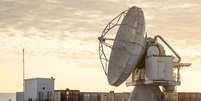 A Base Espacial Pituffik (antiga Base A&eacute;rea de Thule) &eacute; mostrada no norte da Groenl&acirc;ndia, em 4 de outubro de 2023. A imagem mostra uma grande antena parab&oacute;lica em frente a cont&ecirc;ineres, com um c&eacute;u alaranjado e nublado ao fundo. Foto: Thomas Traasdahl/Ritzau Scanpix/AFP via Getty Images / BBC News Brasil