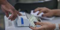 Zoom em um eleitor reconhecendo a digital para votar; em primeiro plano, m&atilde;o de mes&aacute;ria segura t&iacute;tulo de eleitor  Foto: Jo&eacute;dson Alves/Ag&ecirc;ncia Brasil / BBC News Brasil
