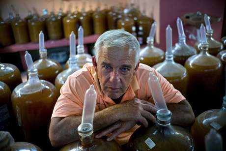 En esta imagen, tomada el 30 de marzo de 2017, Orestes Estévez posa entre docenas de botellas llenas de vino, cerradas con condones, en su casa en La Habana, Cuba. Estévez y su familia llenan los recipientes con jugos elaborados con uvas, guayabas, berros o flor de Jamaica y los cierran colocando un preservativo en el cuello de la botella, para iniciar un inusual proceso de fabricación de vino en una tierra famosa por su ron.