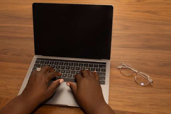 Imagem mostra as mãos de uma pessoa negra digitando no teclado de um Notebook. Ao lado tem um óculos de grau.