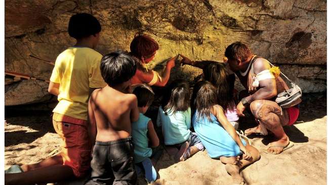 Um índio Waurá ensinando a mitologia em torno do guerreiro Kamukuwaká a crianças da aldeia usando as gravuras da caverna no Xingu antes do local ser vandalizado