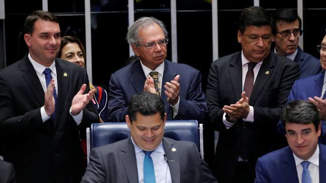 O senador Flávio Bolsonaro, o ministro Paulo Guedes (na foto, os dois à esquerda e em pé) e o presidente da Casa, Davi Alcolumbre (sentado, ao centro), entre outros, comemoram aprovação de texto-base no Senado