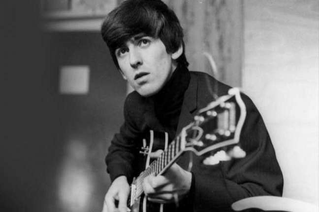 George Harrison de cabelos curtos, sentado e fazendo um bend na guitarra