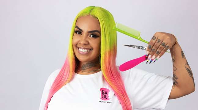 Imagem mostra Esther Gomes, uma mulher com cabelo liso nas cores verdes e rosa. Ela sorri e segura em uma das mãos uma tesoura e dois pentes de cabelo.