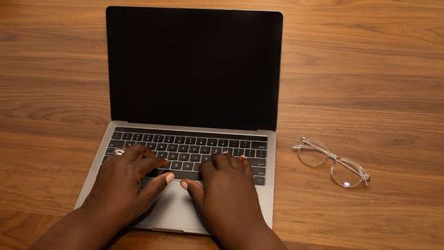Imagem mostra as mãos de uma pessoa negra digitando no teclado de um Notebook. Ao lado tem um óculos de grau.