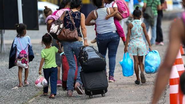 A imagem enquadra três mulheres, sendo duas com uma criança ao colo e outra carregando malas e bolsas. Ao redor delas tem mais três crianças