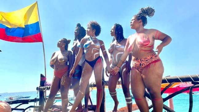 A imagem mostra cinco mulheres negras, do coletivo Birtonga Travel, em viagem à Colombia. Elas vestem biquine, estão na praia e há uma bandeira do país à esquerda