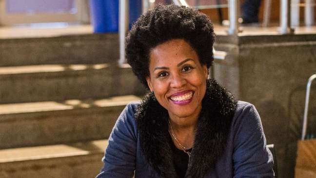 A imagem mostra a escritora Cristiane Sobral. Ela está sentada, veste uma camisa azul e sorri A imagem mostra a escritora Cristiane Sobral. Ela está sentada, veste uma camisa azul e sorri