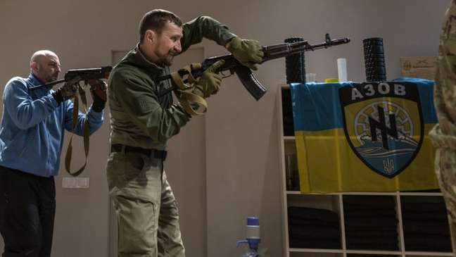 Civilian volunteers are trained by veterans of the Azov Battalion against the Russian invasion;  on the right, the regiment's insignia with variations of two symbols associated with Nazism, the wolf hook and the black sun