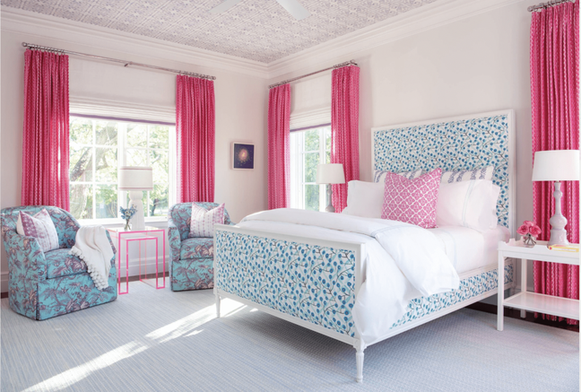 Quarto com cama de casal, poltronas com detalhes azuis, e cortinas rosas.
