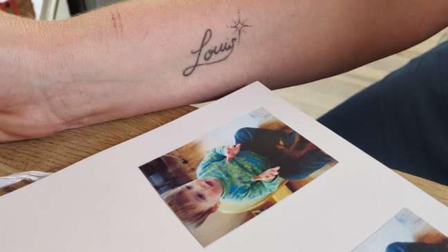 Foto de Louis em álbum de foto e uma tatuagem do seu nome na parte inferior do braço de sua mãe