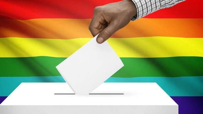 Imagem enquadra uma bandeira LGBT+ ao fundo e uma mão negra que coloca um bilhete em uma urna