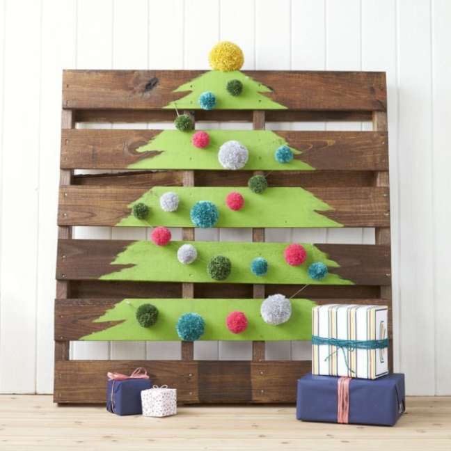 Paint the shape of a tree on wooden pallets and tie pompoms in various colors and sizes to decorate the slats. Cover one of them with gold to imitate a star and voilà , you have a Christmas tree! Paint the shape of a tree on wooden pallets and tie pompoms in various colors and sizes to decorate the slats. Cover one of them with gold to imitate a star and voilà , you have a Christmas tree!