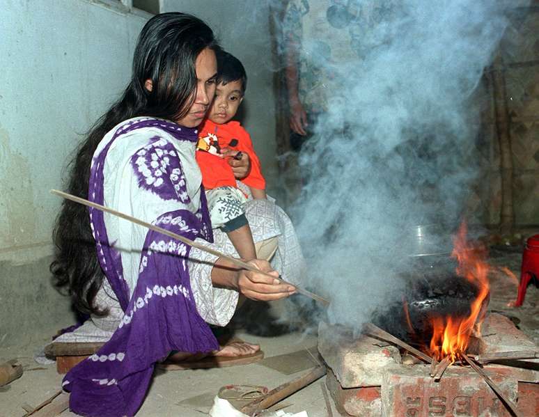 Mulher segura criança enquanto cozinha arroz em fogueira na localidade de Rampura, em Bangladesh