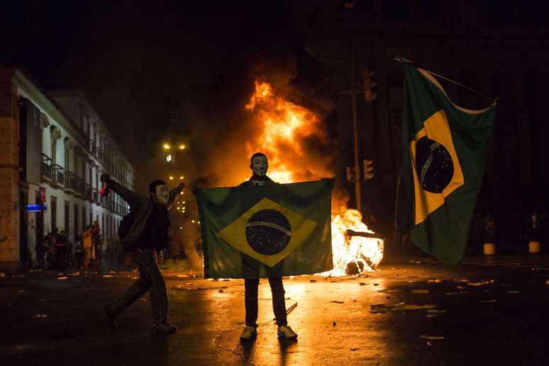 <p>Mascarado, manifestante mostra a bandeira do Brasil em frente a uma barricada de lixo em chamas no Rio</p>