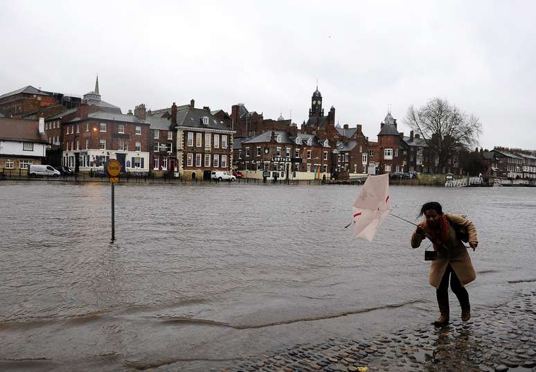 Mulher tenta se proteger do vento e da chuva em calçada alagada pelo rio Ouse em York, na Inglaterra