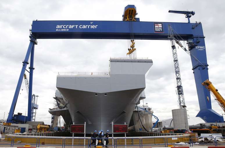 <p>Trabalhadores caminham em frente ao "HMS Queen Elizabeth", o primeiro de dois porta-aviões construídos pela empresa britânica de defesa BAE Systems, no Rosyth Dockyard em Fife, Escócia, em 17 de junho</p>