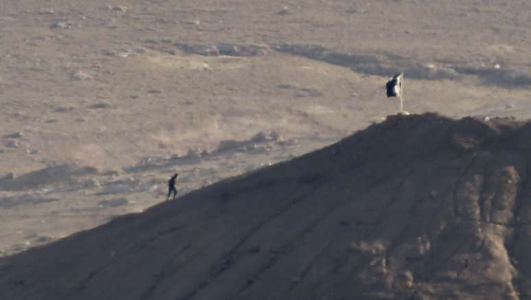 <p>Um militante do&nbsp;Estado Isl&acirc;mico caminha pr&oacute;ximo a uma&nbsp;bandeira do grupo terrorista cravada em um monte&nbsp;perto da cidade s&iacute;ria de Kobani, em&nbsp;6 de outubro de 2014&nbsp;</p>