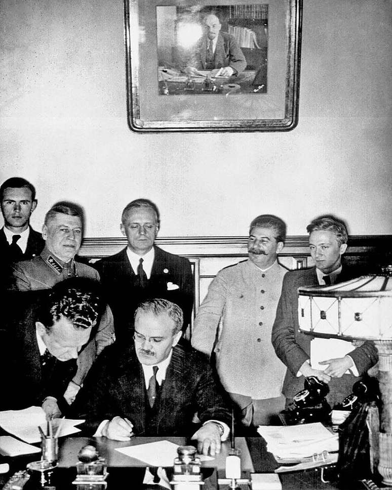 Cerimônia de assinatura do Pacto: imagem mostra Molotov assinando e Ribbentrop atrás (com os olhos fechados), com Stalin à sua esquerda