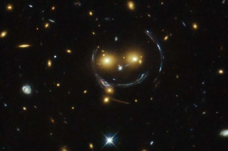 Os olhos do "rosto feliz" são duas galáxias brilhantes distantes, e as linhas que compõem a face e o sorriso arcos criados por um efeito chamado de lente gravitacional forte