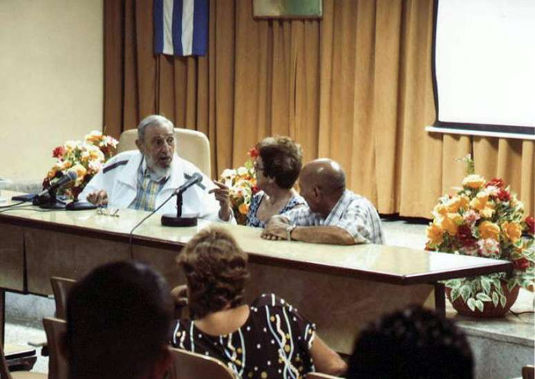 Ex-presidente de Cuba aparece vestido com uma jaqueta branca esportiva e camisa quadriculada, sentado na cabeceira de uma sala de reuniões e acompanhado de dois funcionários cubanos