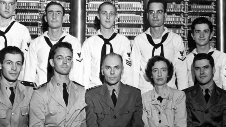 Grace Hopper se juntou &agrave; equipe de cientistas de computa&ccedil;&atilde;o de Harvard liderada pelo comandante Howard Aiken (linha inferior, centro); Hopper foi a primeira programadora do Harvard Mark 1 (em segundo plano na foto), o primeiro computador eletromec&acirc;nico, constru&iacute;do na IBM e enviado para Harvard em 1944, que tinha cerca de 800 quil&ocirc;metros de cabos.