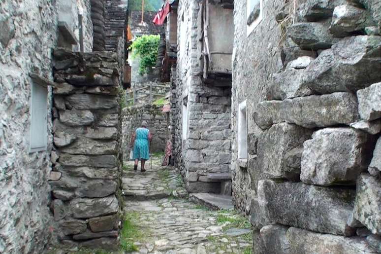 Senhora de vestido azul caminha de bengala por via estreita de Corippo, ladeada por paredes de pedras