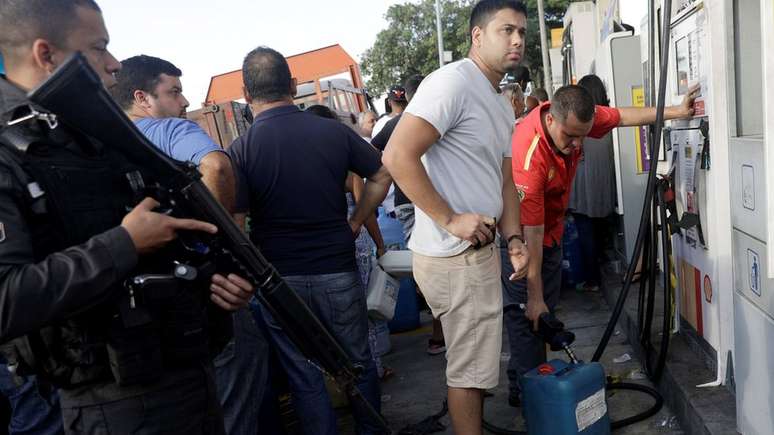 Policial armado patrulha posto de gasolina no Rio de Janeiro, enquanto pessoas enchem galões com combustível