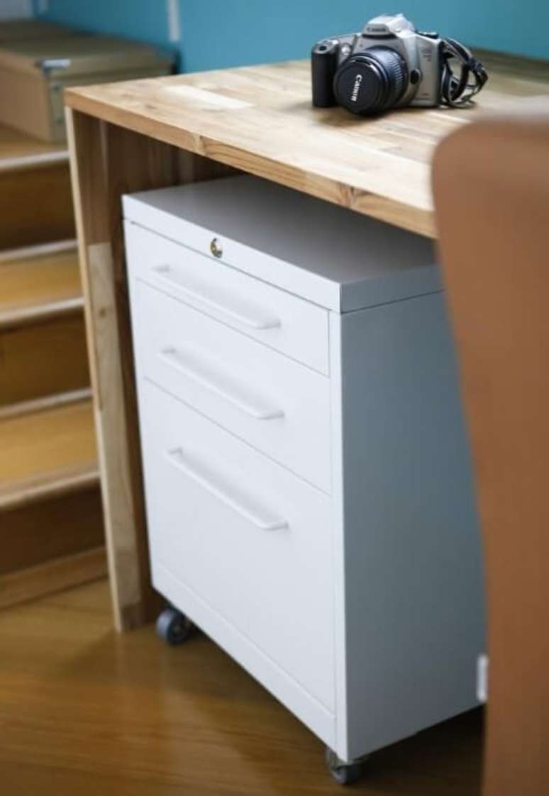 23. Aqui, o gaveteiro aparece embaixo da mesa de madeira usada no home office no quarto. Projeto de Buji