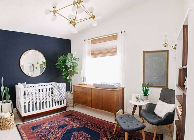 14. Decoração para quarto de bebê com tapete tipo persa, parede azul marinho, espelho redondo e cadeira de amamentação moderna – Foto: Project Nursery