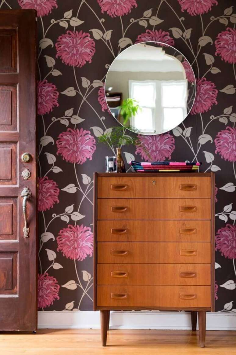 12. Decoração com estilo retrô para ambiente com cômoda de madeira, espelho redondo e papel de parede floral rosa com fundo escuro – Foto: Hunker