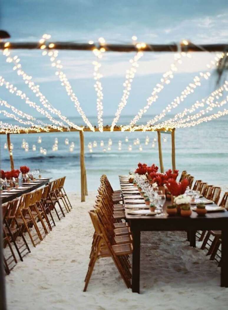 10. Decoração com arranjos de flores vermelhas e varal de luzinhas para mesas de casamento na praia simples – Foto: LM Weddings
