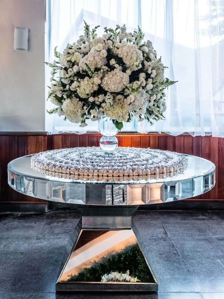 11- A mesa com lembrancinhas de bodas de prata é espelhada e possui um grande vaso com flores brancas. Fonte: Vestida de Noiva