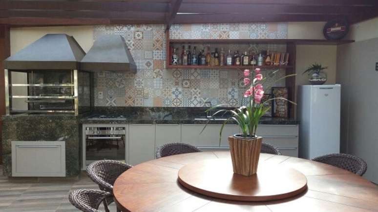 62. &Aacute;rea de lazer aconchegante com churrasqueira de vidro, mesa de madeira redonda e parede revestida com ladrilhos hidr&aacute;ulicos. Fonte: Pinterest