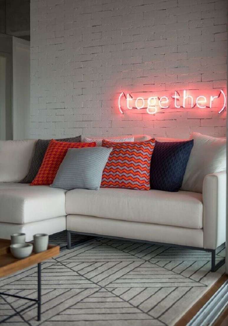 43. Sala moderna decorada com sofá branco, almofadas coloridas e luminária neon em parede de tijolinho branco – Foto: Casa de Valentina