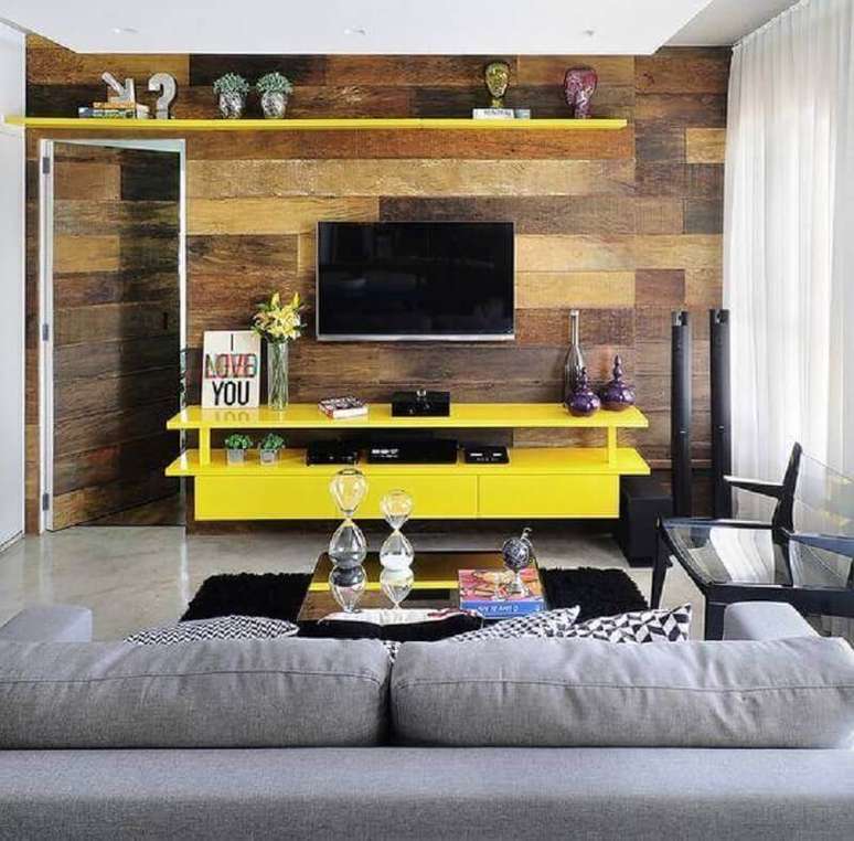 4. Decoração com rack suspenso amarelo para sala decorada com revestimento de madeira e sofá cinza – Foto: Viajando no Apê
