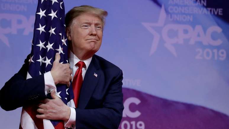 Donald Trump abraça a bandeira americana na reunião anual da CPAC, perto de Washington; Eduardo Bolsonaro repetiu gesto em evento brasileiro