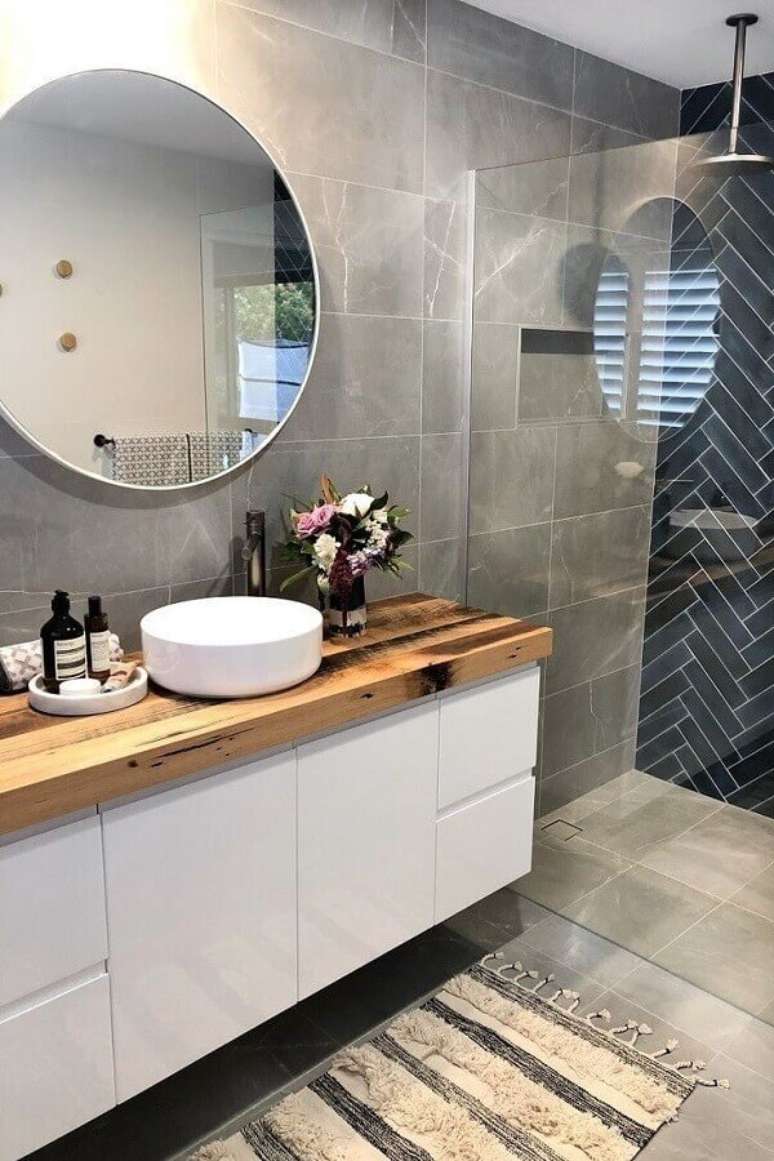 16. Revestimento cinza para banheiro com bancada de madeira e espelho redondo – Foto: Creative Remodeling