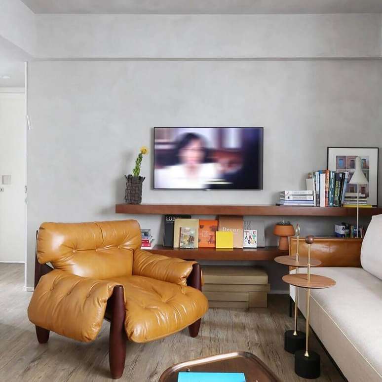 55. Sala com parede cinza claro decorada com prateleiras de madeira e poltrona mole de couro – Foto: MAB3 Arquitetura
